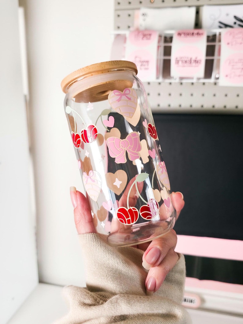 May include: A clear glass tumbler with a wooden lid, decorated with pink bows, hearts, and red cherries. The tumbler is held in a person's hand. The design is printed on the glass. The tumbler is designed for cold beverages.