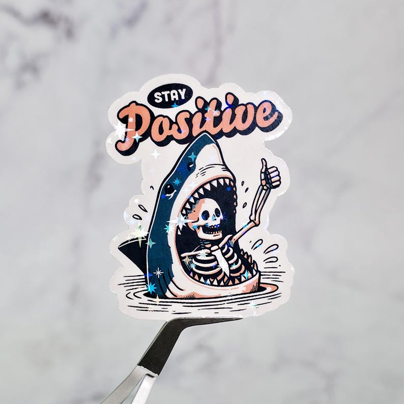 May include: A holographic sticker featuring a cartoon shark with a skeleton inside, giving a thumbs-up. The sticker has the words "Stay Positive" in a pink and orange font with star accents. The shark is blue and white.