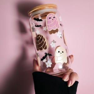 May include: A clear glass can-shaped tumbler with a bamboo lid, decorated with cartoon concha bread designs in pink, brown, and white, along with small white flowers and black bat silhouettes. The tumbler is held against a pink background.