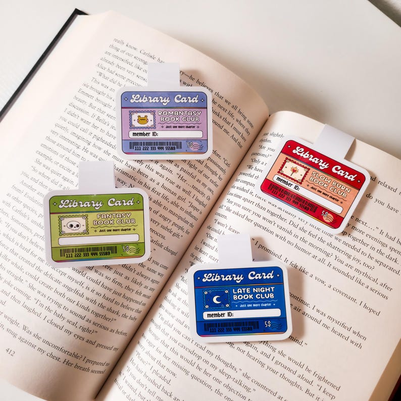May include: Four colorful bookmarks with a library card theme. Each bookmark features a different book club theme: "Romantasy Book Club", "Fantasy Book Club", "Slow Burn Book Club", and "Late Night Book Club". Each bookmark has a cartoon illustration and a member ID number.
