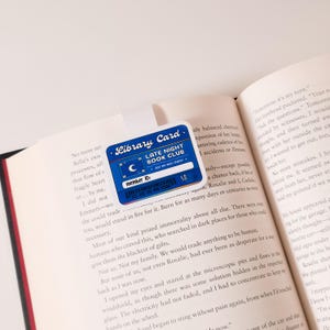 May include: A blue and white library card with the text "Library Card" and "Late Night Book Club". The card has a membership number and a barcode. The card is on top of an open book.