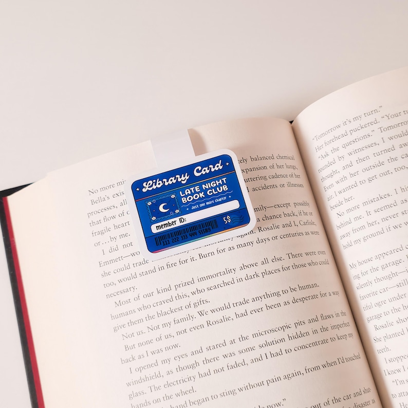 May include: A blue and white library card with the text "Library Card" and "Late Night Book Club". The card has a membership number and a barcode. The card is on top of an open book.
