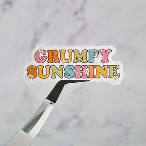 May include: A holographic sticker with the words "GRUMPY SUNSHINE" in colorful, retro-style lettering. The sticker has a white border and small daisy accents. The sticker is being held by a pair of black-tipped tweezers.