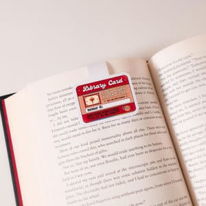 May include: A red and white library card with the text "Slow Burn Book Club" and "Just one more chapter". The card has a small illustration of a tree and the words "member ID:" followed by a series of numbers.