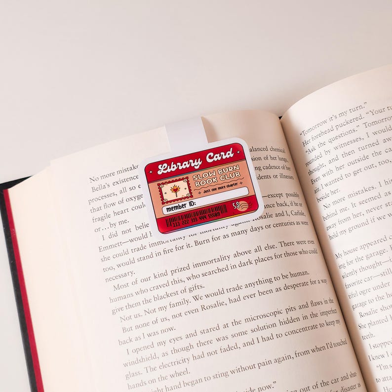 May include: A red and white library card with the text "Slow Burn Book Club" and "Just one more chapter". The card has a small illustration of a tree and the words "member ID:" followed by a series of numbers.