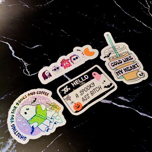 K&ouml;nnte beinhalten: Eine Auswahl an Halloween-Stickern auf einer schwarzen Marmoroberfl&auml;che. Die Aufkleber zeigen Geister, Kaffeetassen und S&auml;tze wie "Cold Like My Heart" und "A Spooky Ass Bitch". Die Aufkleber haben ein holografisches Finish.