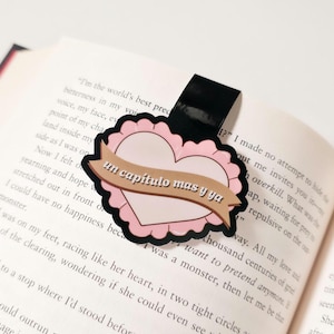 May include: A pink heart-shaped bookmark with a banner that reads "un capitulo mas y ya" in brown text. The bookmark has a black magnetic strip and is placed on the pages of a book.