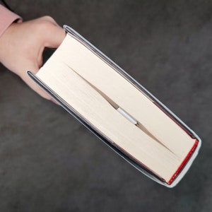 May include: A close-up of a book with a white cover and a black spine. The book is open and a silver bookmark is visible.