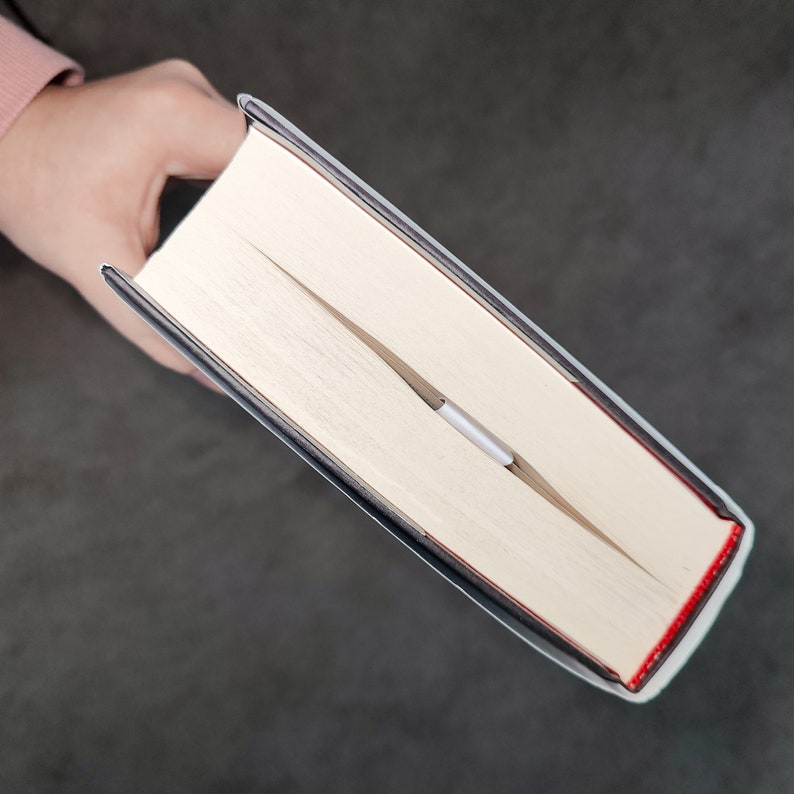 May include: A close-up of a book with a white cover and a black spine. The book is open and a silver bookmark is visible.