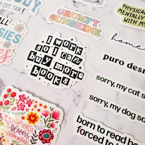 May include: A collection of colorful stickers with various designs and text. Some stickers say "Moody," "Stay Positive," "Grumpy Sunshine," and "I work so I can buy more books." Other stickers feature floral and whimsical illustrations.