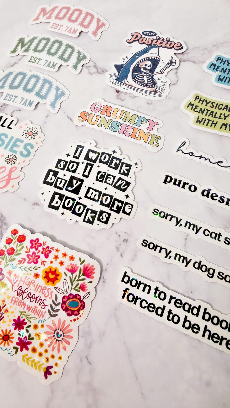 May include: A collection of colorful stickers with various designs and text. Some stickers say "Moody," "Stay Positive," "Grumpy Sunshine," and "I work so I can buy more books." Other stickers feature floral and whimsical illustrations.