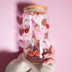 May include: A clear glass tumbler with a bamboo lid, decorated with pink hearts, bows, and cherries. The tumbler is held against a pink background. The cherries are red and the bows are pink.