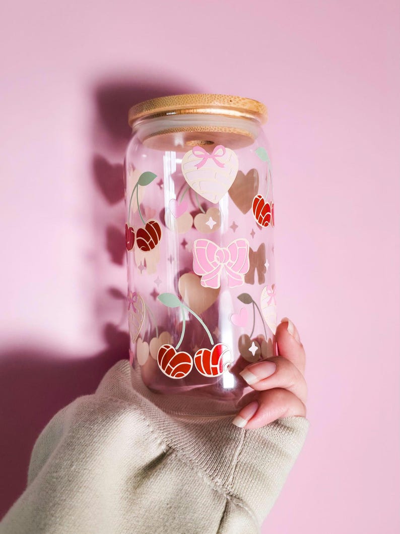 May include: A clear glass tumbler with a bamboo lid, decorated with pink hearts, bows, and cherries. The tumbler is held against a pink background. The cherries are red and the bows are pink.