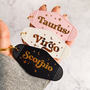 May include: Three keychains shaped like motel room keys with gold lettering on a white background. The keychains are labeled "Taurus", "Virgo", and "Scorpio".