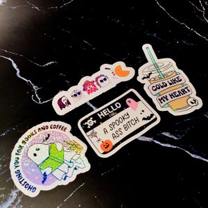 May include: A set of five holographic Halloween stickers featuring ghosts, a skull, a pumpkin, a coffee cup, and a spooky message. The stickers are on a black marble background.