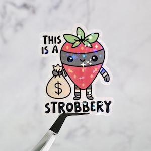 May include: A holographic sticker featuring a cartoon strawberry character with a black mask and striped arms and legs. The strawberry is holding a money bag with a dollar sign. The text reads "THIS IS A STROBBERY."
