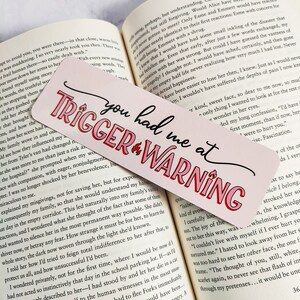 Puede incluir: Marcap&aacute;ginas rosa y rojo con el texto "You had me at Trigger Warning" en una fuente blanca en negrita. El marcap&aacute;ginas tiene un fondo rosa con un gr&aacute;fico de llama roja.