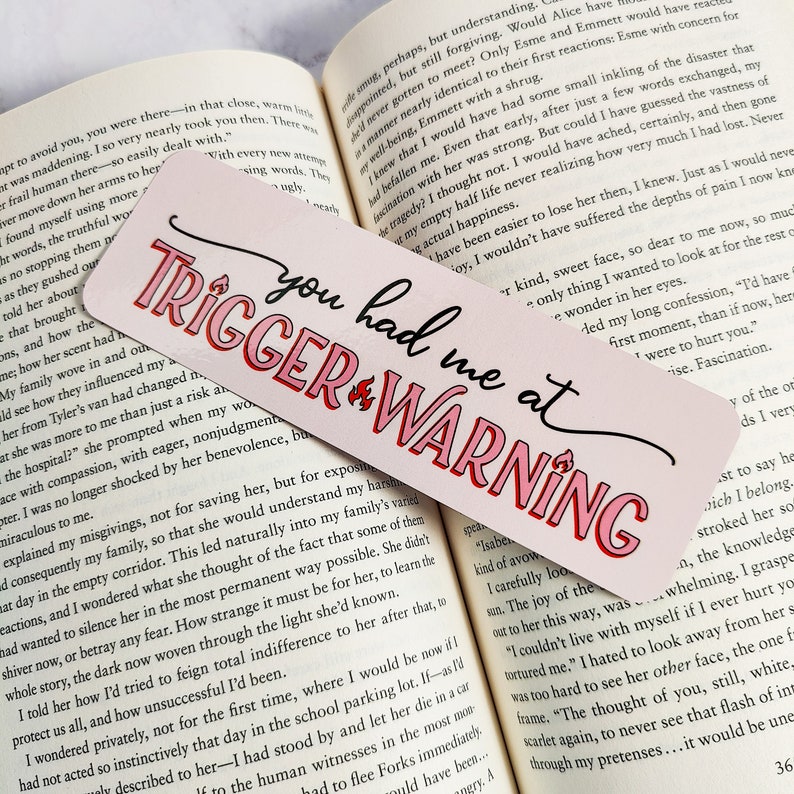 Puede incluir: Marcap&aacute;ginas rosa y rojo con el texto "You had me at Trigger Warning" en una fuente blanca en negrita. El marcap&aacute;ginas tiene un fondo rosa con un gr&aacute;fico de llama roja.