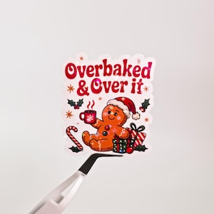May include: A sticker featuring a gingerbread man wearing a Santa hat, holding a mug, and surrounded by Christmas gifts and holly. The sticker has the text "Overbaked & Over it" in red and pink.