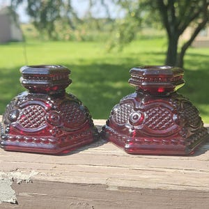 May include: A pair of deep red glass candle holders featuring a pressed pattern with diamond-shaped textures, circular accents, and a beaded border along the base. Each piece has a square-shaped top opening and a wide, stable base design.