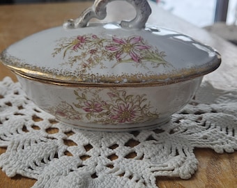 Könnte beinhalten: Eine weiße Porzellanschale mit Deckel, verziert mit einem zarten Blumenmuster in Rosa und Grün. Die Schale und der Deckel sind mit einem abgenutzten Goldrand versehen. Ein geschwungener, verzierter Griff befindet sich auf dem Deckel.