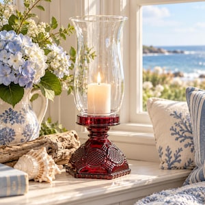 May include: A red glass candle holder with a textured, geometric pattern on the base and a clear glass hurricane shade. A white pillar candle sits inside the holder, emitting a warm, glowing flame. The base has a hexagonal shape with embossed details.