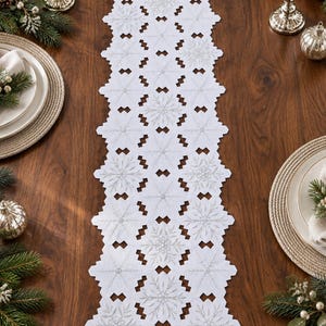 May include: A white table runner featuring a repeating snowflake pattern with intricate silver embroidery. The runner has a die-cut edge design composed of interconnected hexagonal shapes, creating a decorative border along the entire length.