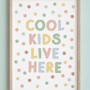 May include: A framed print with the words "COOL KIDS LIVE HERE" in colorful, playful lettering. The background is white with scattered polka dots in pastel shades of pink, yellow, green, and brown. The frame is a light wood color.