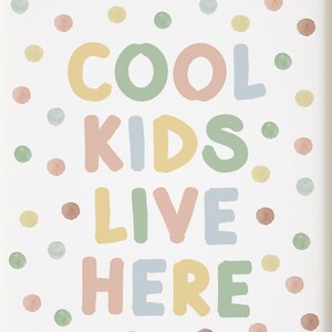 May include: A framed print with the words "COOL KIDS LIVE HERE" in colorful, rounded letters. The text is surrounded by watercolor-style polka dots in pastel shades of yellow, green, pink, and blue. The frame is a light wood color.