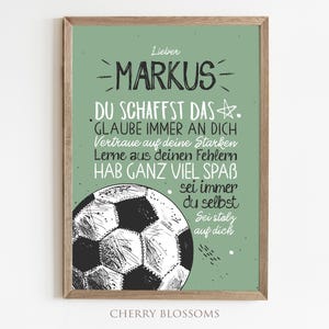 May include: A framed print with a green background and a soccer ball illustration. The print features the name "Markus" and inspirational text in German. The frame is a light brown color. The words "Cherry Blossoms" are at the bottom.