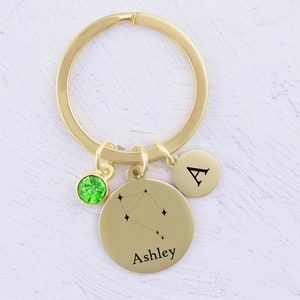 May include: Gold keychain with a green crystal charm, a round charm with the letter "A" and a larger round charm with a constellation design and the name "Ashley" engraved on it.