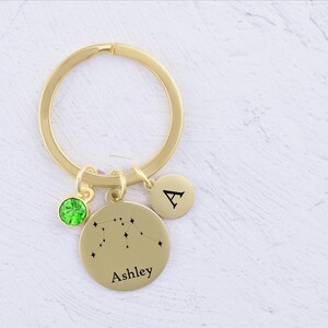 May include: Gold-tone keychain with a green crystal charm, a round charm with a constellation design and the name "Ashley", and a round charm with the letter "A".