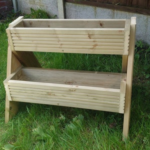 May include: A two-tiered wooden planter box with a natural finish. The rectangular boxes are constructed with horizontal slats and supported by angled legs. The planter is set on a grassy lawn, ideal for gardening.