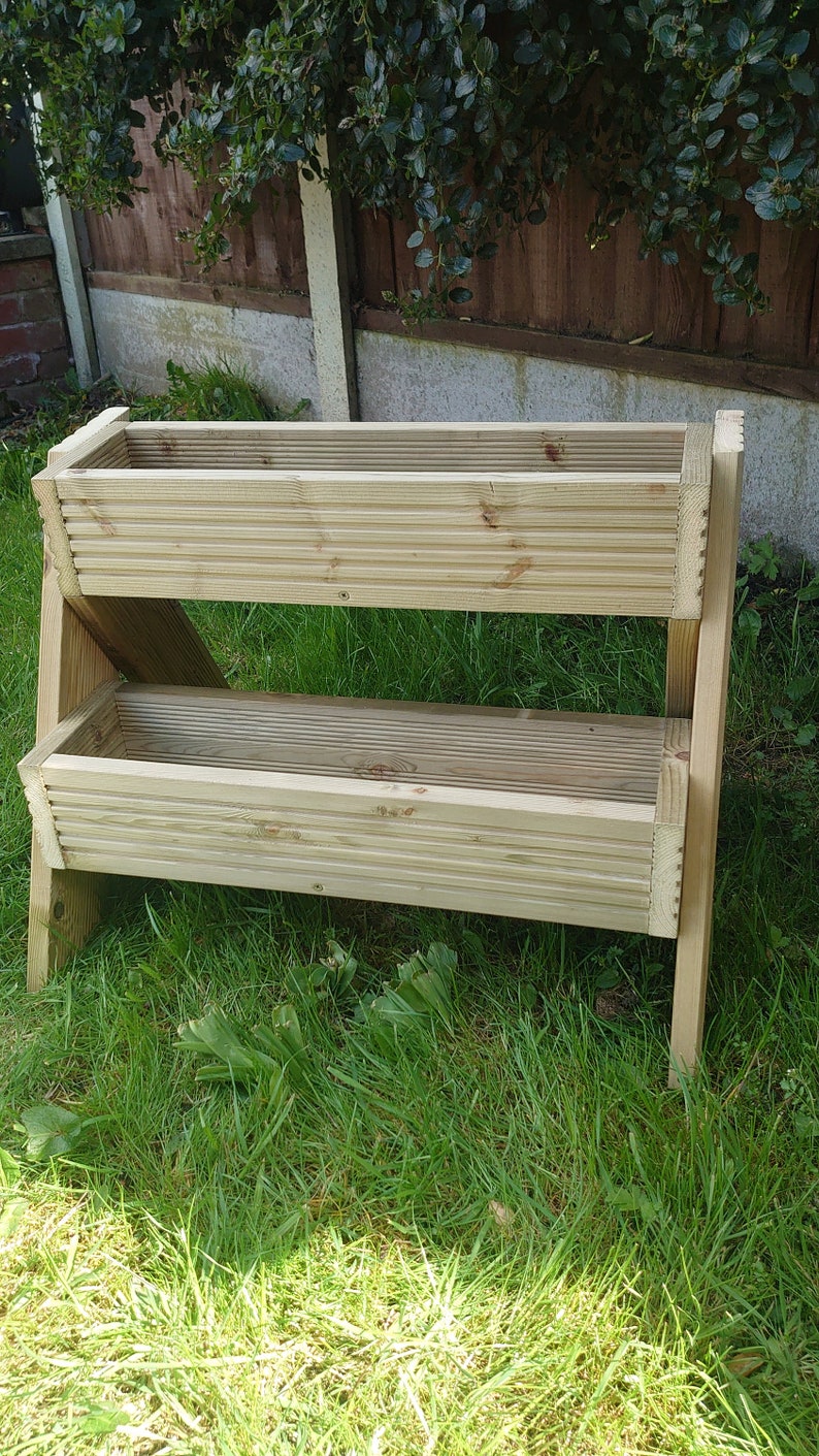 May include: A two-tiered wooden planter box with a natural finish. The rectangular boxes are constructed with horizontal slats and supported by angled legs. The planter is set on a grassy lawn, ideal for gardening.