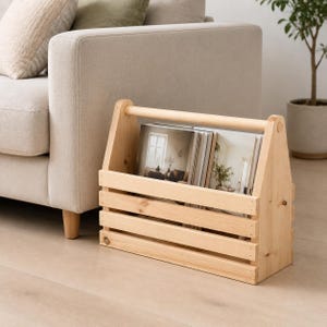 May include: A light-coloured wooden magazine rack with a slatted design and a top handle. The rack is constructed from natural pine wood and contains several magazines with visible interior photography, showcasing a clean and organised storage solution.