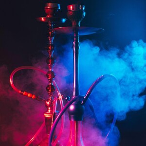 Op de afbeelding: Twee waterpijpen met rode en blauwe accenten tegen een donkere achtergrond, met de roze tekst "HOOKAH LOUNGE". De afbeelding is gevuld met kleurrijke rook, wat een levendige en sfeervolle scène creëert.