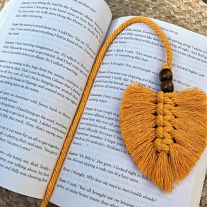 May include: Mustard yellow macrame bookmark with a heart-shaped design and a tassel. The bookmark is lying on an open book, with the text of the book visible. The bookmark is made of a woven, knotted material.