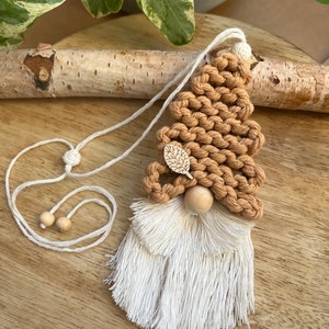 May include: A brown macrame gnome ornament with a white tassel beard and a wooden nose. The gnome is hanging from a white cord with wooden beads.