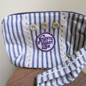 May include: A blue and white striped fabric pouch with a white scalloped trim. The pouch has a zipper closure and a long strap. It features three white buttons and a purple patch with the text "Atlantic Coast Line".