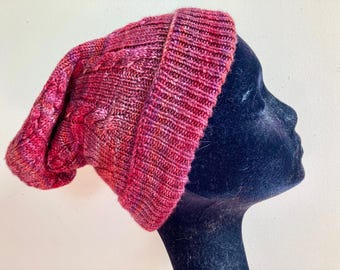 May include: A hand-knitted beanie in shades of red and pink, featuring a cable knit design. The hat is displayed on a black mannequin head against a white background. The beanie has a folded brim and a slouchy fit.