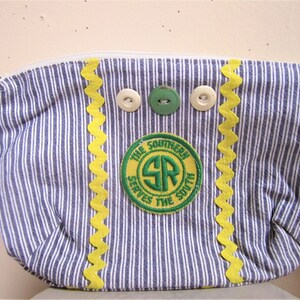 May include: A blue and white striped fabric pouch with a yellow zig-zag trim. The pouch has three buttons and a green and yellow patch that reads "The Southern Serves The South" with the letters "SR" in a circle.