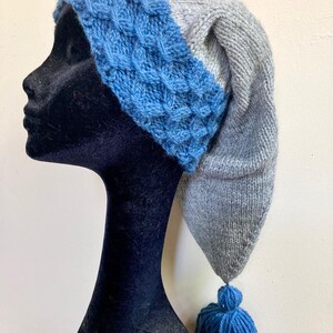 May include: A knitted hat with a long, pointed end and a tassel. The hat features a blue textured band and a gray body. The tassel and band are blue. The hat is displayed on a black mannequin head.