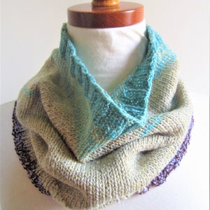 May include: A knitted cowl with a teal blue and beige color pattern. The cowl is folded over and has a purple and teal blue trim.