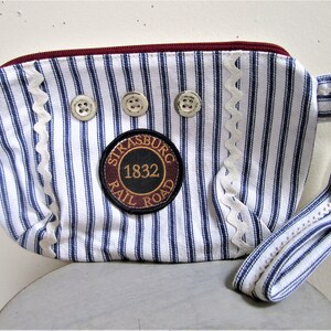May include: A blue and white striped fabric pouch with a zipper closure and a handle. The pouch has a white lace trim and three silver buttons. A brown circular patch with the text "Strasburg Rail Road 1832" is sewn on the front.