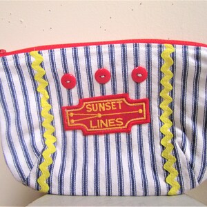 May include: A blue and white striped fabric zippered pouch with a yellow zig-zag trim and red buttons. The pouch has an embroidered patch that says "SUNSET LINES" in yellow letters on a red background.