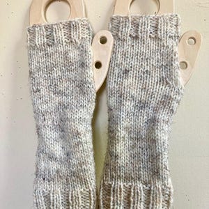 May include: A pair of hand-knitted fingerless gloves in a speckled, light gray yarn. The gloves are displayed on wooden forms with a circular top and two holes. The cuffs and tops of the gloves have a ribbed design.