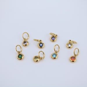 K&ouml;nnte beinhalten: Eine Sammlung goldfarbener Anh&auml;nger mit verschiedenfarbigen Edelsteinen. Die Anh&auml;nger haben oben eine kleine Schlaufe zum Befestigen an einer Kette oder einem Armband. Zu den Edelsteinfarben geh&ouml;ren Gr&uuml;n, Lila, Blau, Gelb, Klar und Rot.