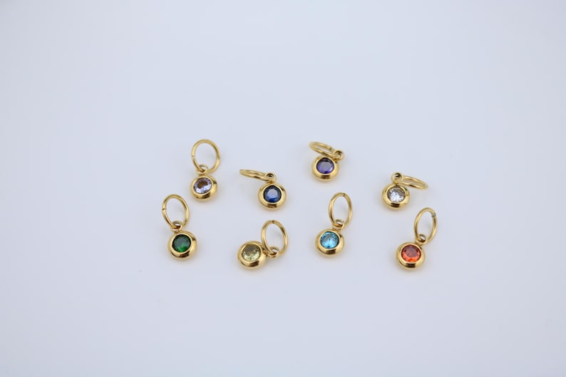 K&ouml;nnte beinhalten: Eine Sammlung goldfarbener Anh&auml;nger mit verschiedenfarbigen Edelsteinen. Die Anh&auml;nger haben oben eine kleine Schlaufe zum Befestigen an einer Kette oder einem Armband. Zu den Edelsteinfarben geh&ouml;ren Gr&uuml;n, Lila, Blau, Gelb, Klar und Rot.