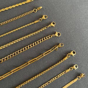 K&ouml;nnte beinhalten: Eine Auswahl goldfarbener Kettenhalsketten mit verschiedenen Gliederstilen, darunter Seil-, Curb- und B&uuml;roklammer-Designs. Jede Halskette verf&uuml;gt &uuml;ber einen Karabinerverschluss. Die Ketten werden auf grauem Hintergrund pr&auml;sentiert.