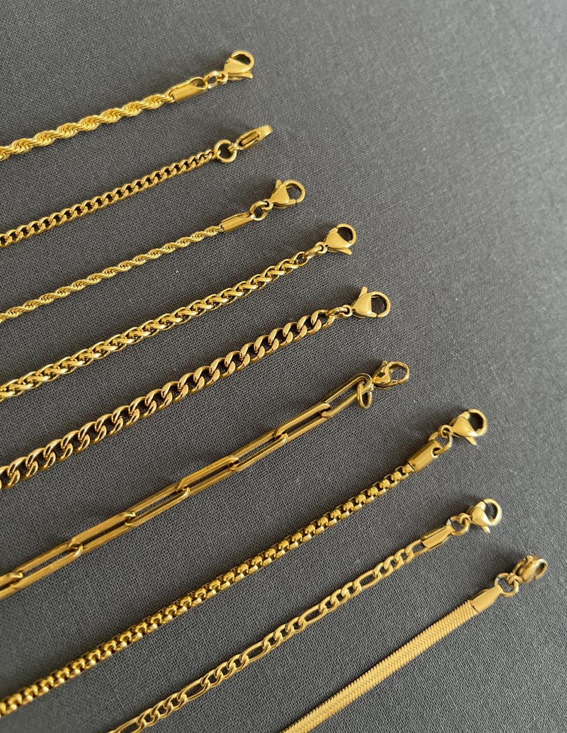 K&ouml;nnte beinhalten: Eine Auswahl goldfarbener Kettenhalsketten mit verschiedenen Gliederstilen, darunter Seil-, Curb- und B&uuml;roklammer-Designs. Jede Halskette verf&uuml;gt &uuml;ber einen Karabinerverschluss. Die Ketten werden auf grauem Hintergrund pr&auml;sentiert.